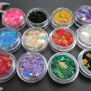 May include: Twelve small jars filled with iridescent flower-shaped glitter in various colors, including pink, blue, black, gold, white, red, purple, and green.