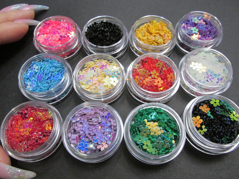 May include: Twelve small jars filled with iridescent flower-shaped glitter in various colors, including pink, blue, black, gold, white, red, purple, and green.