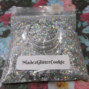 Puede incluir: Una bolsa de plástico transparente y un pequeño recipiente llenos de purpurina plateada. La purpurina tiene una calidad reflectante e iridiscente. La etiqueta de la bolsa dice "Made2GlitterCookie". El fondo presenta un estampado floral.