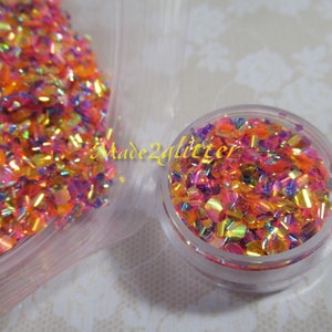 May include: A close-up of a small jar filled with iridescent glitter in shades of pink, orange, and gold. The glitter is in a variety of shapes and sizes, and it is very sparkly. The jar is sitting on a white surface. Made2glitter is written on the jar.