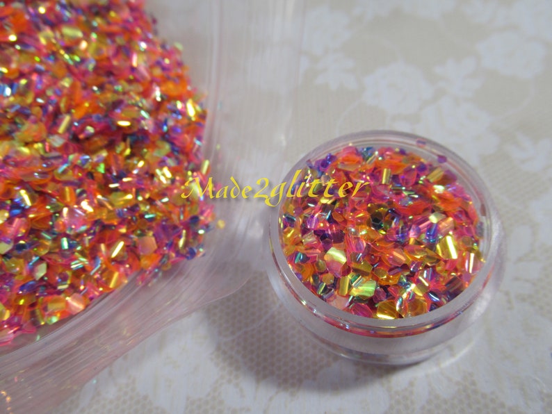 May include: A close-up of a small jar filled with iridescent glitter in shades of pink, orange, and gold. The glitter is in a variety of shapes and sizes, and it is very sparkly. The jar is sitting on a white surface. Made2glitter is written on the jar.