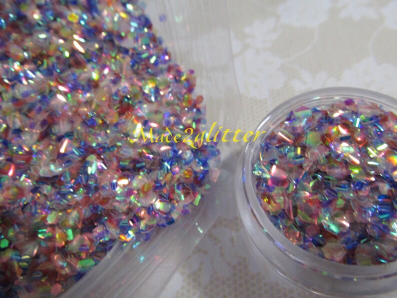 May include: A close-up of iridescent glitter in a clear plastic container. The glitter is a mix of pink, blue, and green colors. The glitter is in a small container and a larger container. The text "Made2glitter" is visible on the larger container.