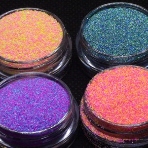 Può includere: Quattro piccoli barattoli di glitter, ognuno con un colore diverso: rosa, viola, verde acqua e giallo. I barattoli sono disposti a forma di quadrato su una superficie nera.