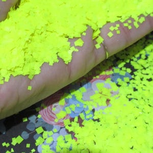 Puede incluir: Una mano sosteniendo un montón de confeti cuadrado amarillo neón brillante. El confeti está esparcido sobre una superficie con estampado floral. El confeti es pequeño y parece estar hecho de un material fino.