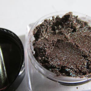 May include: A small jar of dark brown eyeshadow with a shimmery finish. The eyeshadow is in a clear jar with a black lid.