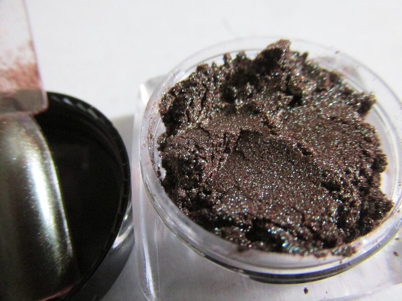 May include: A small jar of dark brown eyeshadow with a shimmery finish. The eyeshadow is in a clear jar with a black lid.