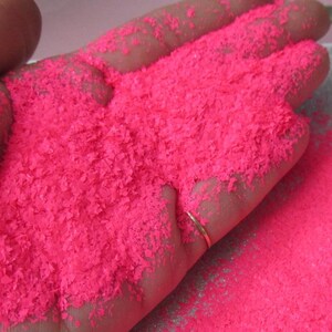 May include: A close-up shot of a hand filled with bright pink, shredded material. The material appears to be finely cut and textured, resembling small flakes. A gold ring is visible on the ring finger. The overall effect is a vibrant display of color and texture.