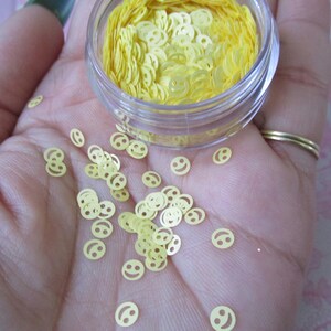 Op de afbeelding: Een klein, doorzichtig potje gevuld met gele smiley-glitter. De glitter is verspreid over een hand.