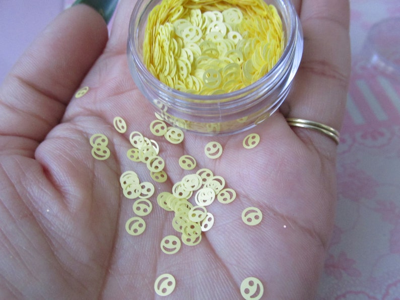 Op de afbeelding: Een klein, doorzichtig potje gevuld met gele smiley-glitter. De glitter is verspreid over een hand.