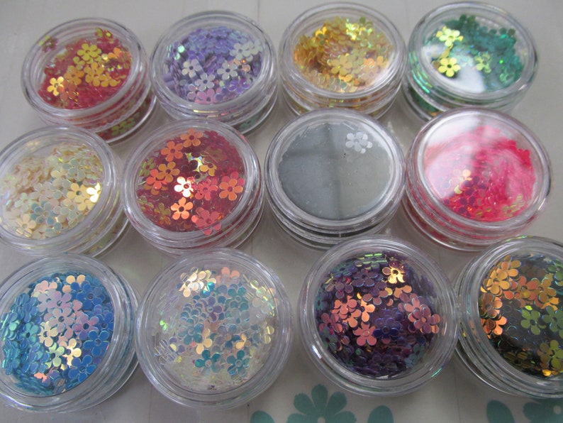 May include: Twelve small jars filled with iridescent flower-shaped glitter. The jars are clear plastic and the glitter is in various colors, including red, pink, green, blue, purple, gold, and silver.