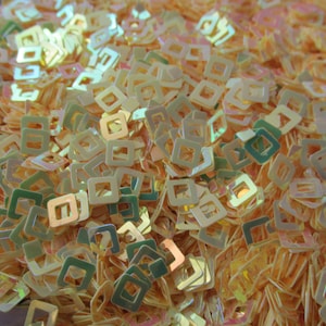 May include: A pile of iridescent, light gold square sequins with a hole in the center. The sequins are scattered randomly and are all the same size.