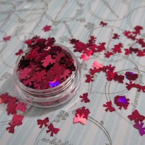 May include: A small clear jar filled with red glitter shaped like bows. The glitter is scattered on a light blue background with a pattern of circles.