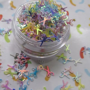 May include: A clear jar filled with iridescent rainbow colored glitter shaped like scissors. The glitter is scattered around the jar on a white background with a colorful pattern.