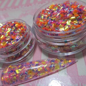 May include: Two clear glass jars filled with iridescent orange, pink, and purple glitter. The glitter is in a variety of shapes and sizes. A clear glass stick with glitter on it is also shown.