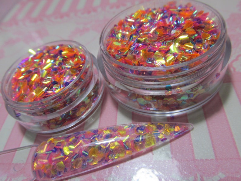 May include: Two clear glass jars filled with iridescent orange, pink, and purple glitter. The glitter is in a variety of shapes and sizes. A clear glass stick with glitter on it is also shown.
