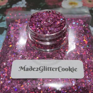 May include: A clear jar filled with iridescent, magenta-colored glitter sits atop a clear plastic bag filled with the same glitter. A white label with the text "Made2GlitterCookie" is on the bag.