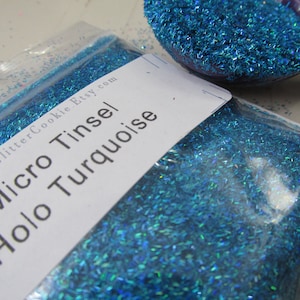 May include: A clear plastic bag filled with turquoise holographic micro tinsel glitter. A white label on the bag reads Made2GlitterCookie.Etsy.com Micro Tinsel Holo Turquoise. A purple plastic scoop filled with the same blue glitter sits nearby.