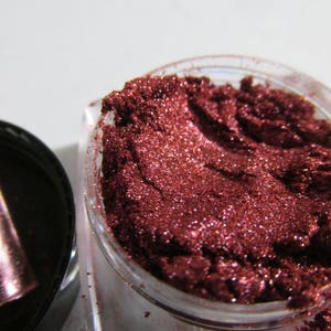 May include: A close-up of a small jar filled with a deep red, glittery makeup pigment.