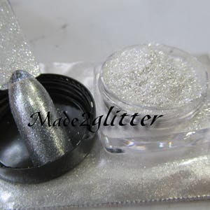 May include: A clear jar filled with silver glitter. The glitter is loose and has a fine texture. The jar is sitting on a white surface. The text "Made2glitter" is visible on the jar.