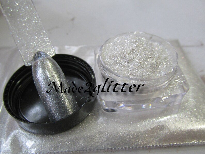 May include: A clear jar filled with silver glitter. The glitter is loose and has a fine texture. The jar is sitting on a white surface. The text "Made2glitter" is visible on the jar.