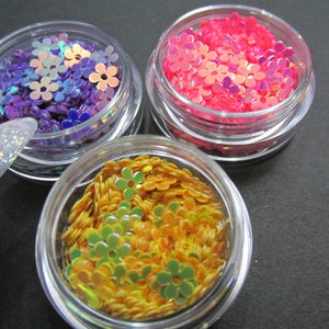 May include: Three small jars filled with iridescent flower-shaped sequins. The jars are clear plastic and the sequins are purple, pink, and orange.