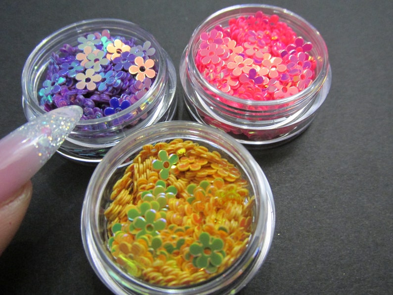 May include: Three small jars filled with iridescent flower-shaped sequins. The jars are clear plastic and the sequins are purple, pink, and orange.