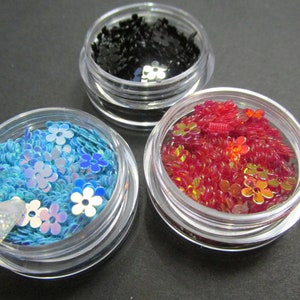 May include: Three small jars filled with colorful glitter and sequins. The jars are clear plastic with silver lids. The glitter is in shades of blue, red, and black.