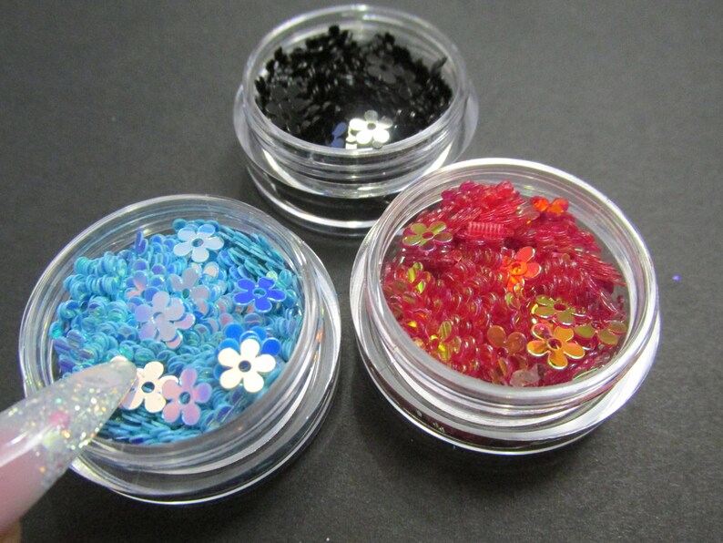 May include: Three small jars filled with colorful glitter and sequins. The jars are clear plastic with silver lids. The glitter is in shades of blue, red, and black.