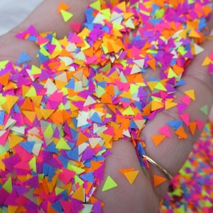 May include: A hand holding a pile of colorful confetti. The confetti is made up of small, triangular pieces in bright neon colors, including pink, orange, yellow, and blue. The confetti is scattered across the hand.