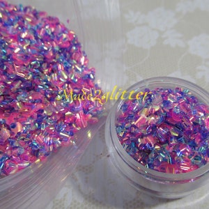 May include: A small clear jar filled with iridescent pink and blue glitter. The glitter is in a variety of shapes and sizes. The jar is sitting on a white surface.