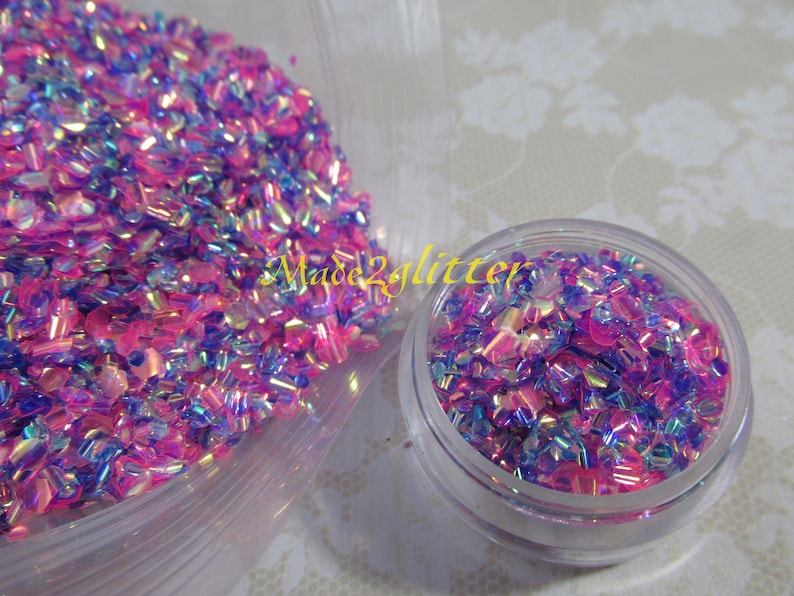 May include: A small clear jar filled with iridescent pink and blue glitter. The glitter is in a variety of shapes and sizes. The jar is sitting on a white surface.
