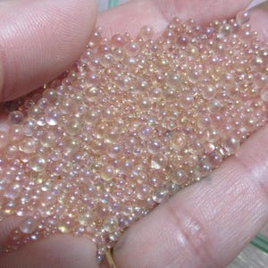 May include: A handful of iridescent, light peach-colored glass beads. The beads vary in size and reflect light, creating a shimmering effect. The beads are small and round, and appear to be loose. The overall effect is delicate and visually appealing.