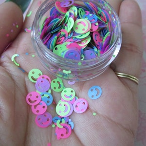 Op de afbeelding: Een klein, doorzichtig potje gevuld met kleurrijk smiley-confetti. Het confetti is in tinten roze, groen, blauw en paars.