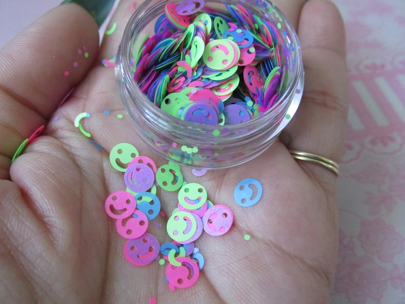 Op de afbeelding: Een klein, doorzichtig potje gevuld met kleurrijk smiley-confetti. Het confetti is in tinten roze, groen, blauw en paars.