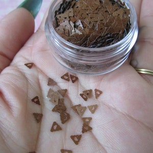 Op de afbeelding: Een klein, doorzichtig potje gevuld met bruine, driehoekige glitter. De glitter is verspreid over een hand.