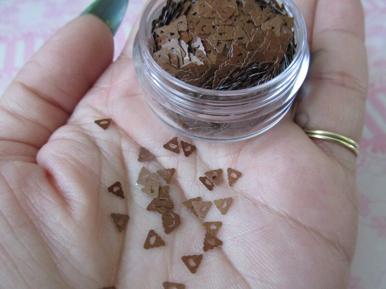 Op de afbeelding: Een klein, doorzichtig potje gevuld met bruine, driehoekige glitter. De glitter is verspreid over een hand.