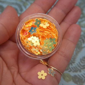 May include: A small clear plastic container filled with bright orange flower-shaped sequins, accompanied by several loose gold-toned metallic flower-shaped sequins resting on the skin, each featuring a central hole and five rounded petals.