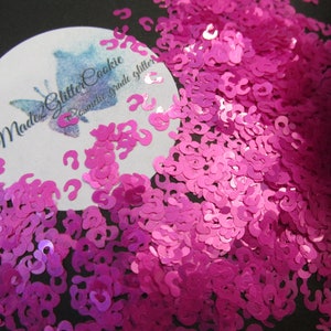 May include: Pink question mark shaped glitter. The glitter is in a pile and is on a black background. The glitter is labeled "Made 2 Glitter Cookie Cosmetic Grade Glitter".