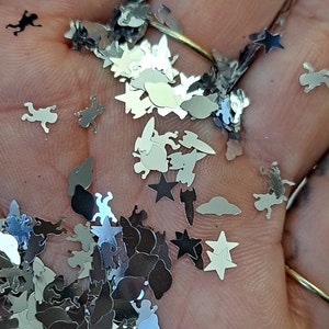 May include: Silver glitter in various shapes, including stars, clouds, rockets, and whimsical figures. The glitter is scattered across a hand, creating a sparkling effect. Ideal for crafts and decorations.