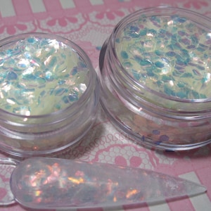 May include: Two clear glass jars filled with iridescent white and blue glitter. A clear plastic spatula with glitter on it is in the foreground.