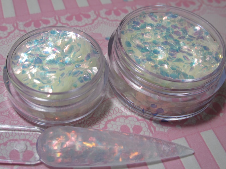 May include: Two clear glass jars filled with iridescent white and blue glitter. A clear plastic spatula with glitter on it is in the foreground.