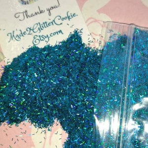 Op de afbeelding: Een klein doorzichtig plastic zakje gevuld met teal-kleurige, iriserende glitter. Het zakje ligt op een roze en wit gepatroneerd oppervlak. De tekst "Made2GlitterCookie Etsy.com" is gedrukt op een witte kaart.