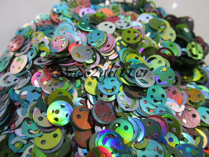 Op de afbeelding: Een close-up afbeelding van iriserende groene, blauwe en paarse glitter in de vorm van smiley's. De glitter is perfect voor knutselen en het toevoegen van glitter aan projecten.