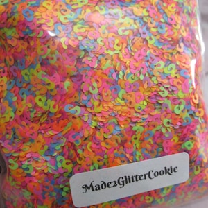 Puede incluir: Una bolsa transparente llena de confeti de colores neón en varias formas. El confeti es principalmente rosa, naranja, amarillo y azul. La etiqueta de la bolsa dice "Made2GlitterCookie".