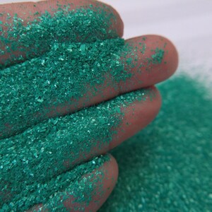 May include: Close-up of a hand covered in sparkling, teal-colored glitter. The glitter is finely ground and reflects light, creating a shimmering effect. The background is a plain white surface, highlighting the glitter's vibrant color and texture. This glitter is ideal for crafts and decorations.