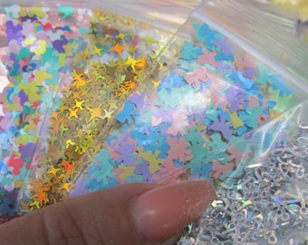 May include: Close-up of several clear plastic bags filled with colorful glitter shapes. The shapes include hearts, stars, butterflies, and scissors. The glitter colors are pink, gold, blue, and iridescent.