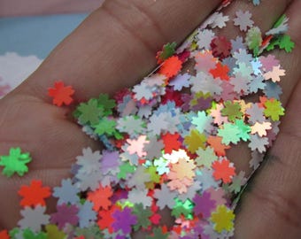 May include: A handful of colorful, flower-shaped sequins in shades of orange, green, pink, purple, and white. The sequins are small and glittery, reflecting light. These decorative embellishments are ideal for crafts and DIY projects.