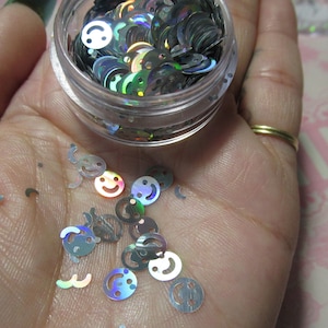 Op de afbeelding: Een klein, doorzichtig potje gevuld met iriserende zilveren glitter in de vorm van smiley's. Sommige glitter is verspreid over een hand.