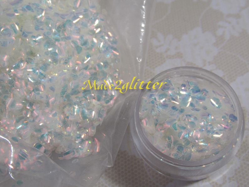 May include: A close-up of iridescent white glitter in a clear plastic bag and a clear jar. The glitter is small and has a holographic finish. The text "Made2glitter" is visible on the bag.