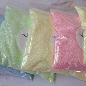 May include: Six small bags of iridescent glitter in various pastel colors, including light blue, green, yellow, pink, and white. Each bag has a small white label with a dragonfly logo and the word "Made".